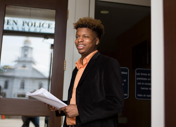 A male student in a suit jacket stands in front of the Hamden Police Department door holding a stack of papers.