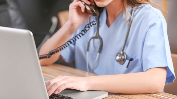 A nurse in scrubs talks on the phone while she types at a computer