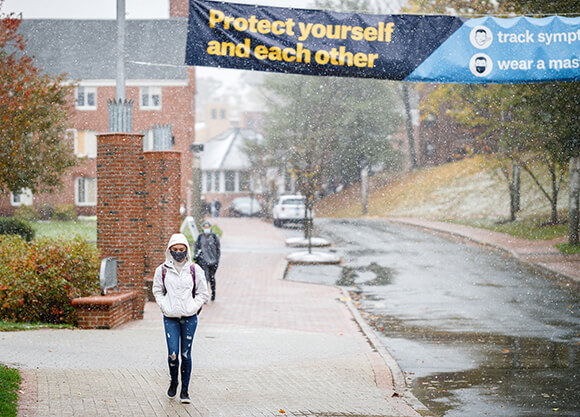 Students walk under a banner reading "Protect Yourself and Others" in the snow.