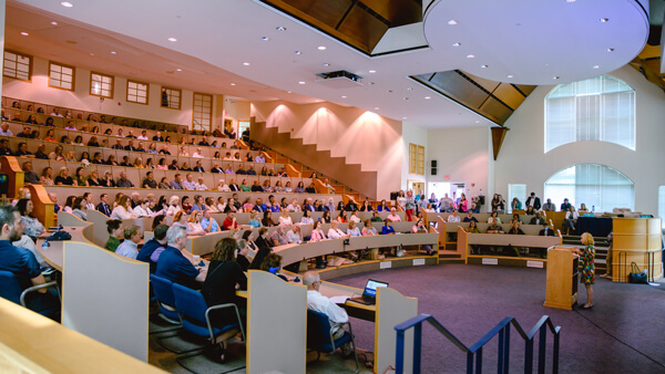 Hundreds of people sit in a large auditorium while a person presents at a podium