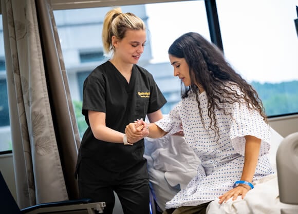 A female student in black scrubs helps a middle aged female patient out of bed