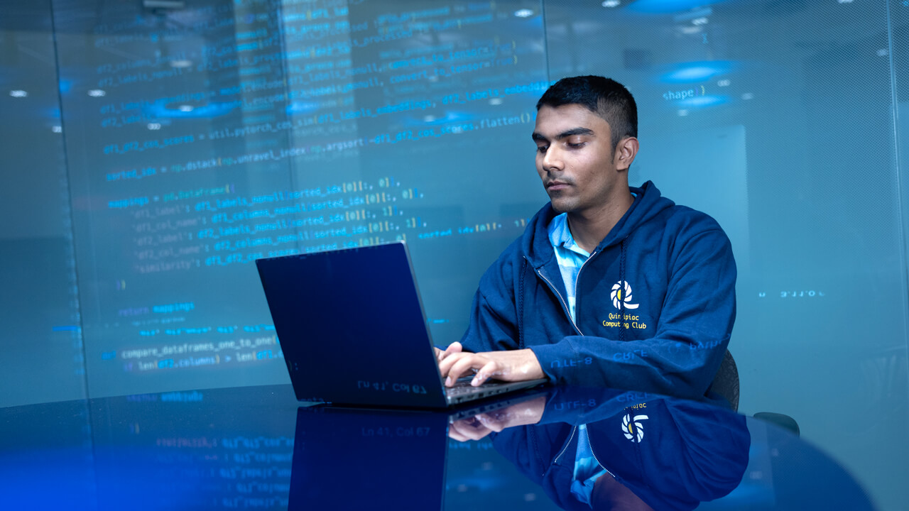Student sitting down and working on a laptop with computer code projected on the back wall