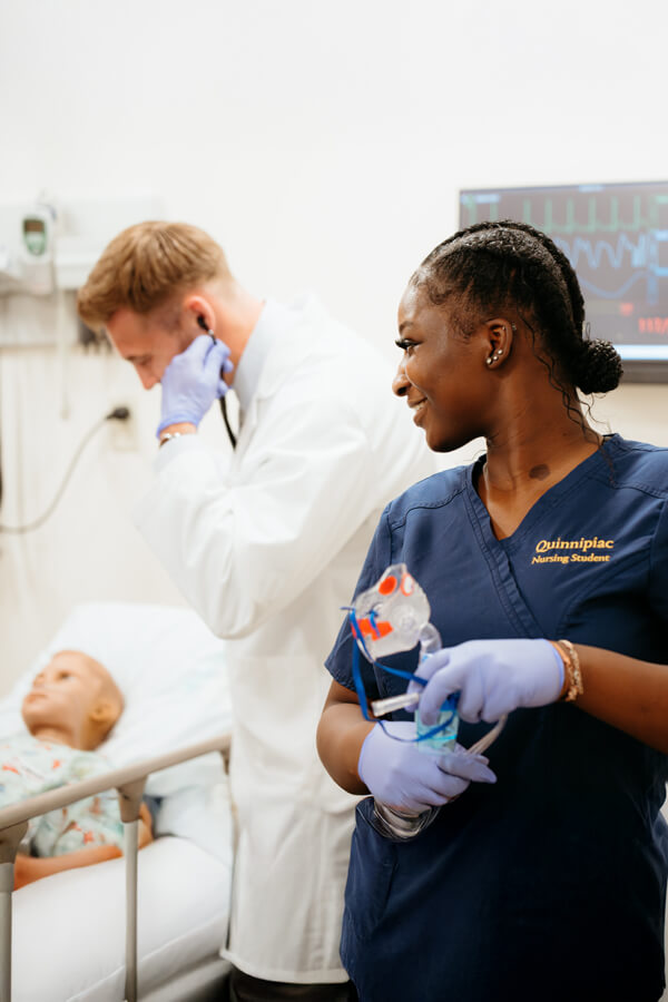 A nursing student smiles while carrying medical equipment through the simulation lab.