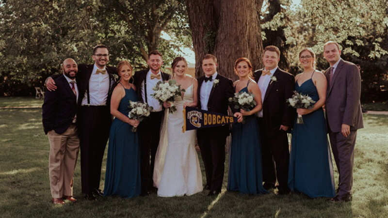 Smiling wedding guests pose under a tree and hold up a Bobcats pennant