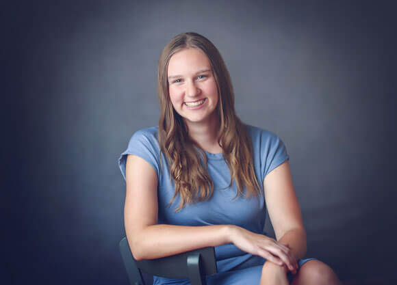 Emma Corridan sitting in a chair smiling against a navy blue background