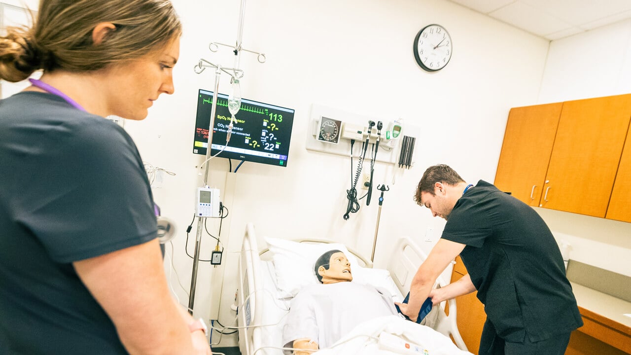 A male student puts a BP cuff on a mannikin while a female student observes