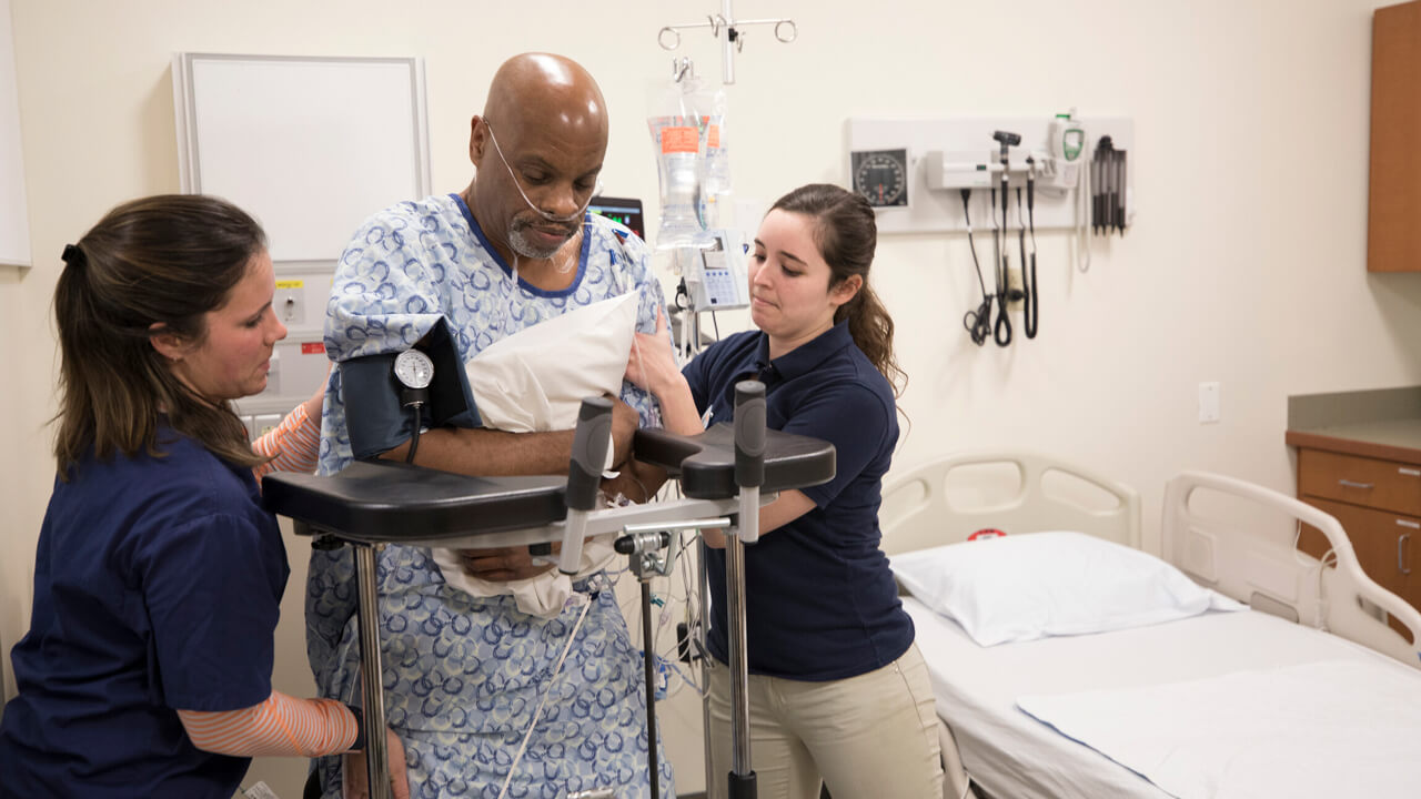 2 female students assist a middle aged man hooked up to oxygen and a BP monitor stand up in a stander