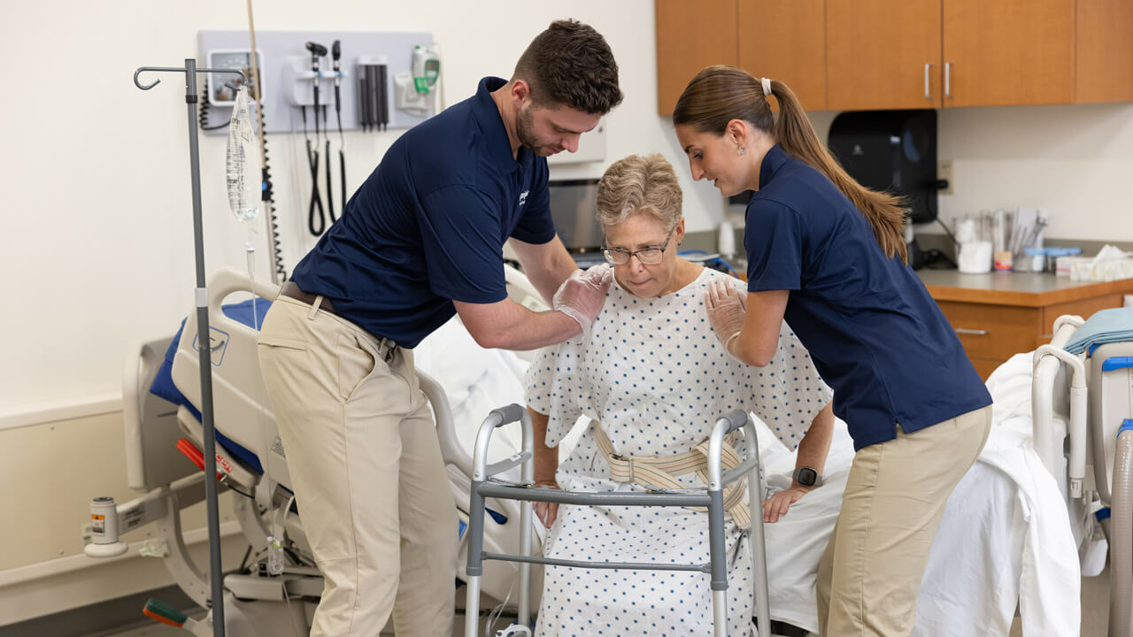 A male and female student help an older woman stand from a hospital bed with a walker