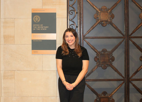 Student smiles for a photo in front of a sign that reads the Office of the Connecticut Secretary of the State