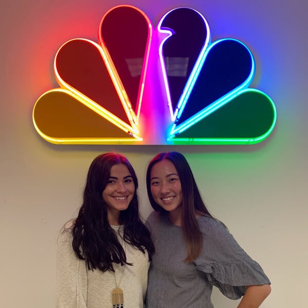 Two student interns posing in front of a sign of the NBC logo