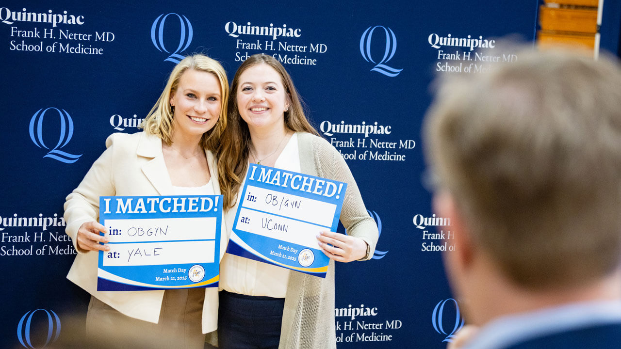 Two School of Medicine students excitingly hold up an 'I Matched!' sign standing against a School of Medicine backdrop