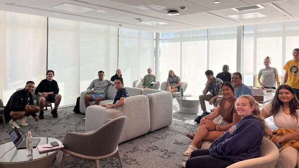 A large group of students smiling at the camera sitting around a room