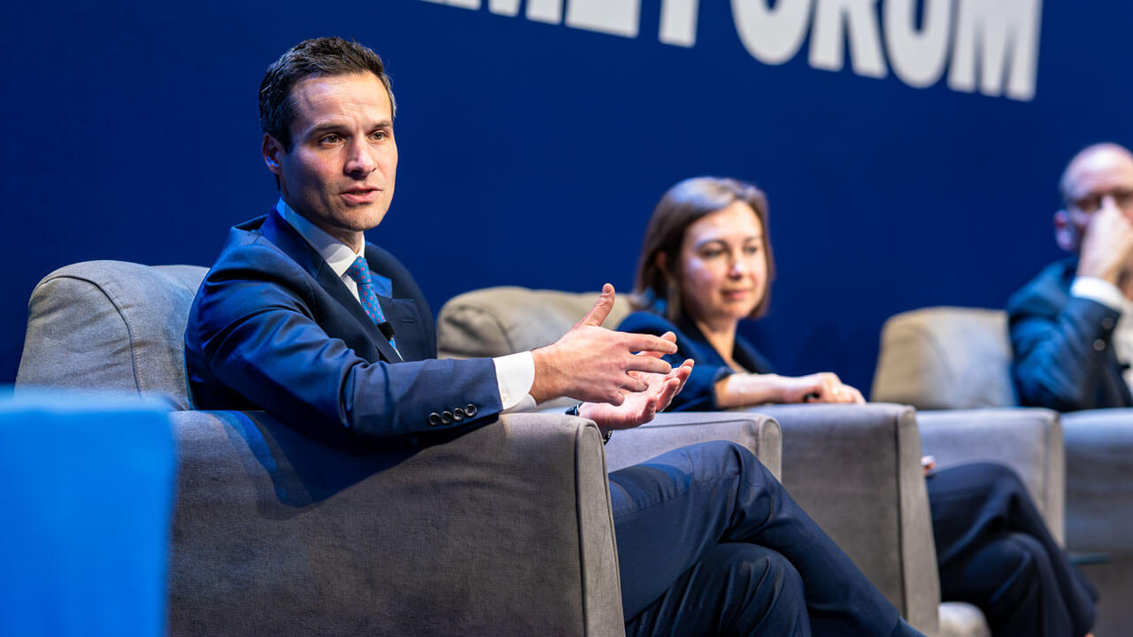 Panelists speak on a stage while sitting in chairs.