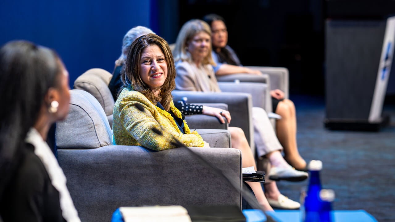 Panelists speak on a stage while sitting in chairs.