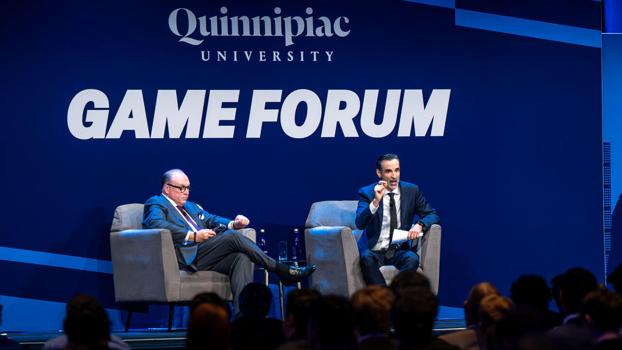 Two individuals sit in chairs on stage and look out into the crowd.