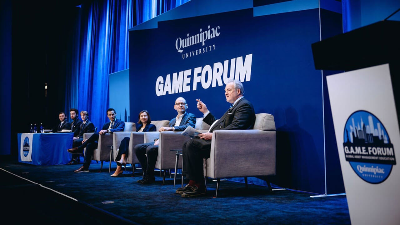 Panelists speak on the stage while sitting in a line of chairs.