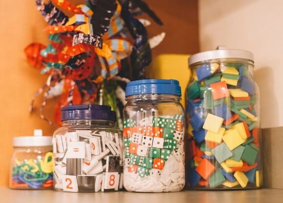Jars of elementary teaching materials sit on a counter, including dice, large paper clips, and numbered and colored tiles.