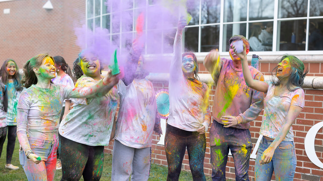 Group of students line up together to throw color powder in the air in front of the student center