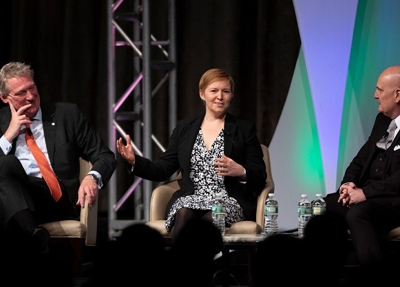 A woman sits in a chair on a stage between two men talking to an audience.