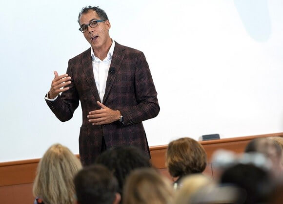 A man stands and speaks in front of a room of people facing him