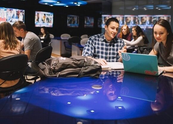 Students work on laptops in front of television screens