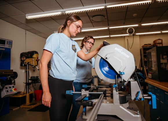 Two school of engineering students using a saw while wearing protective gear
