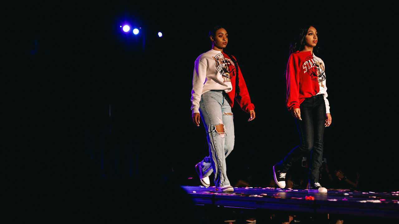 Two students in half red half cream colored sweatshirts walk the runway.