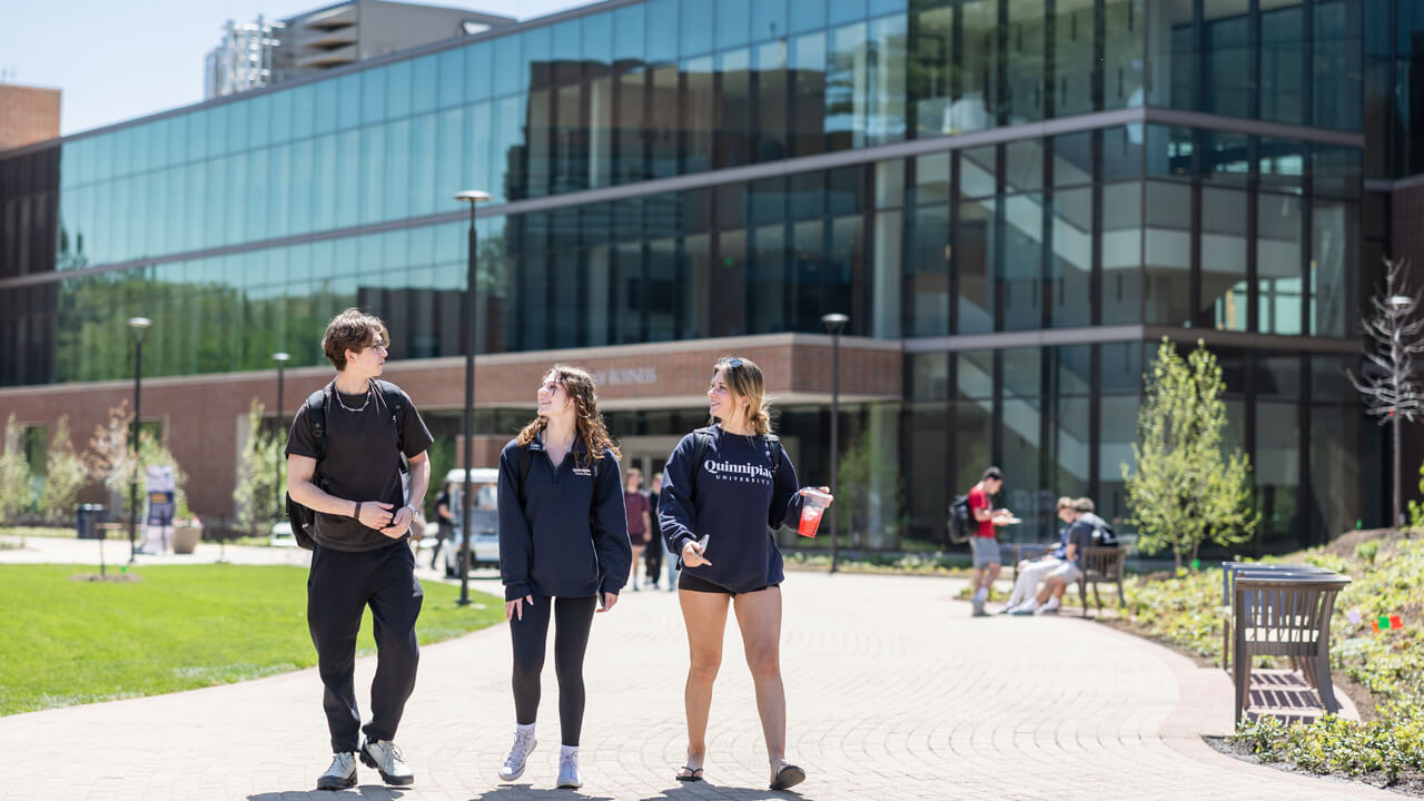 Three students walk along a path in front of the Quinnipiac School of Business on a sunny day