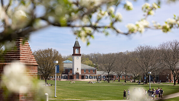 The Arnold Bernhard Library seen through seen through flowering trees as people walk by on the Mount Carmel Campus Quad.
