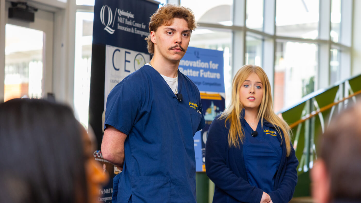 Two Quinnipiac students in navy blue scrubs stand at the front of a room presenting their business idea to a panel of judges during the pitch competition.