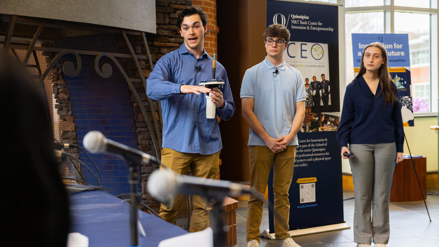 A student explains a prototype device while two team members stand beside him during their pitch presentation in front of a panel of judges.