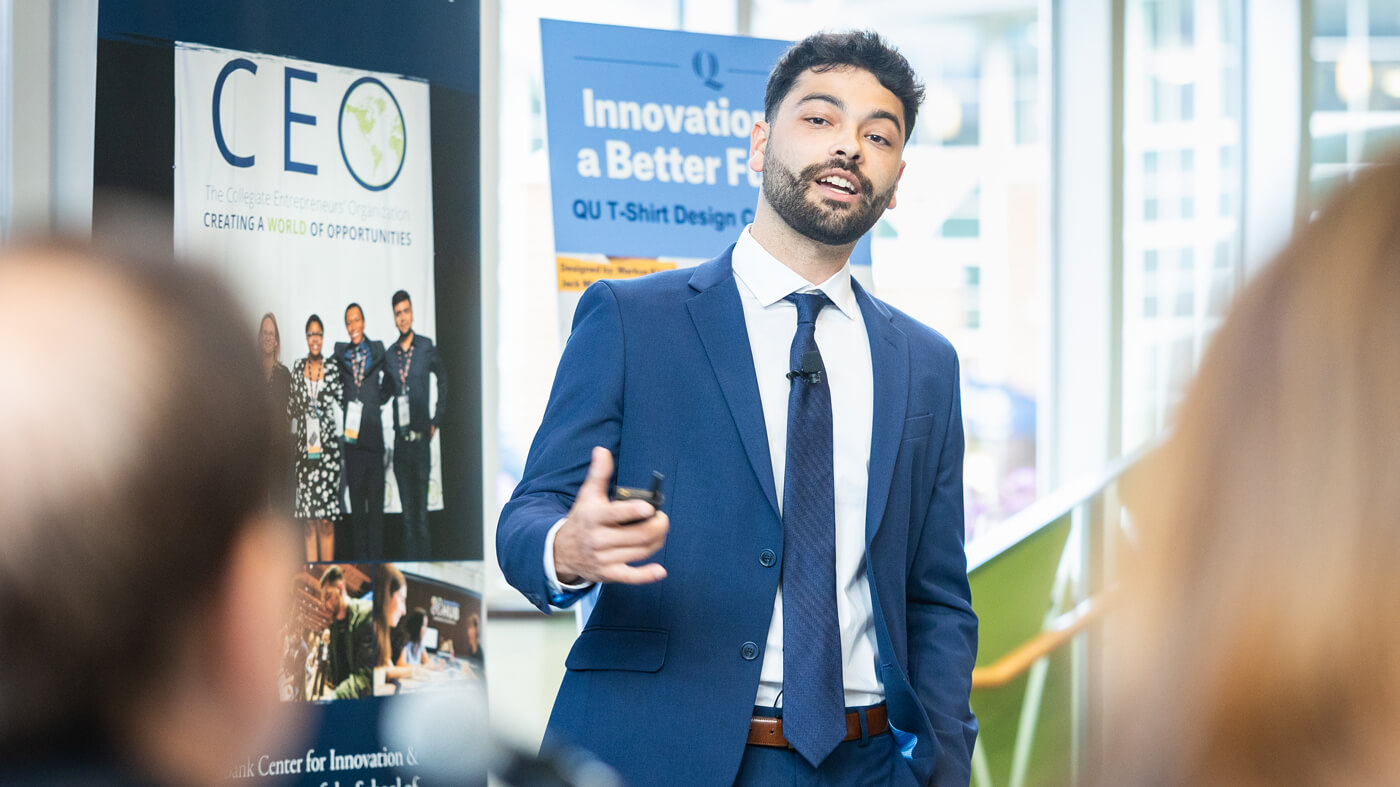 A student in a navy suit gives a confident solo pitch presentation, gesturing with his hand while addressing the judges and audience.