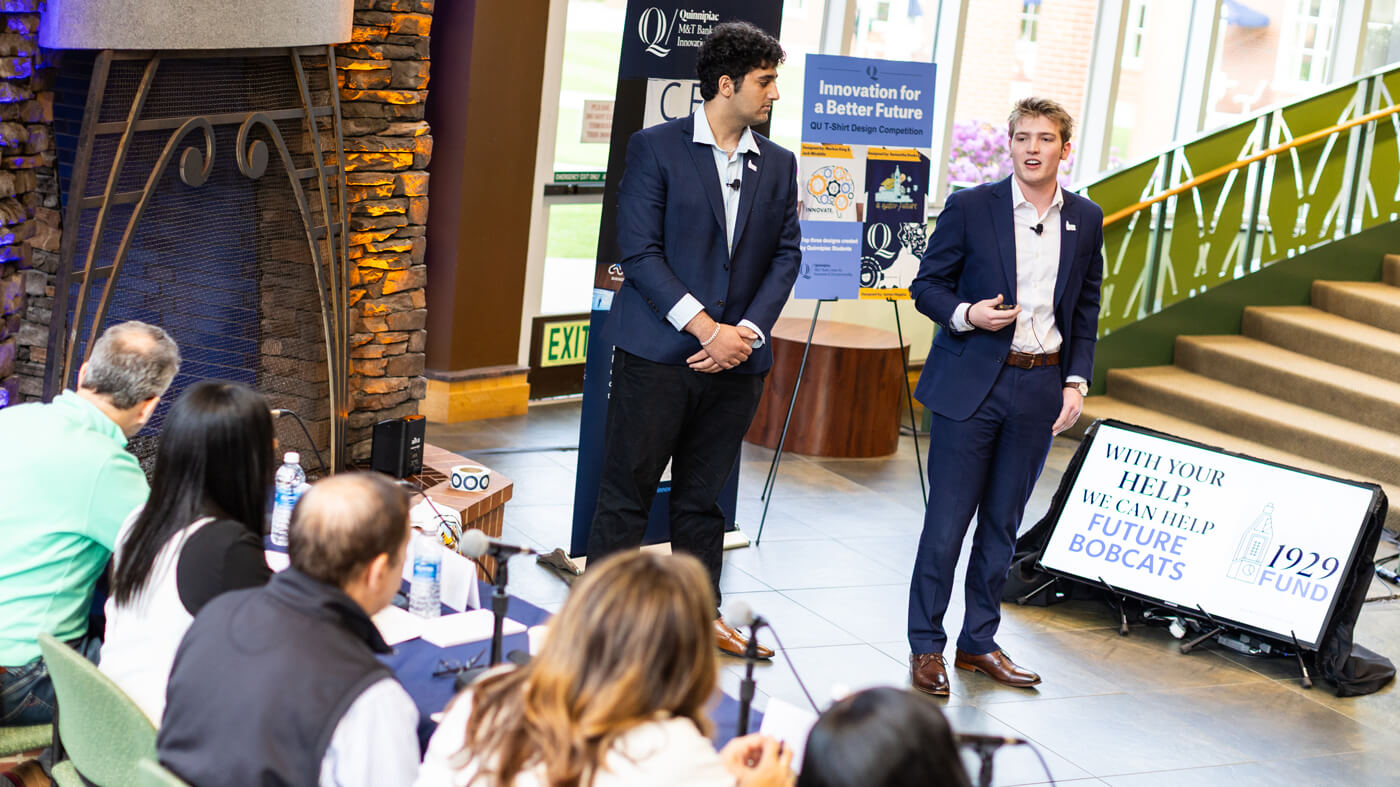 Two students in business attire stand and present their pitch in front of a large screen and a panel of judges, with branding for the 1929 Fund visible beside them.