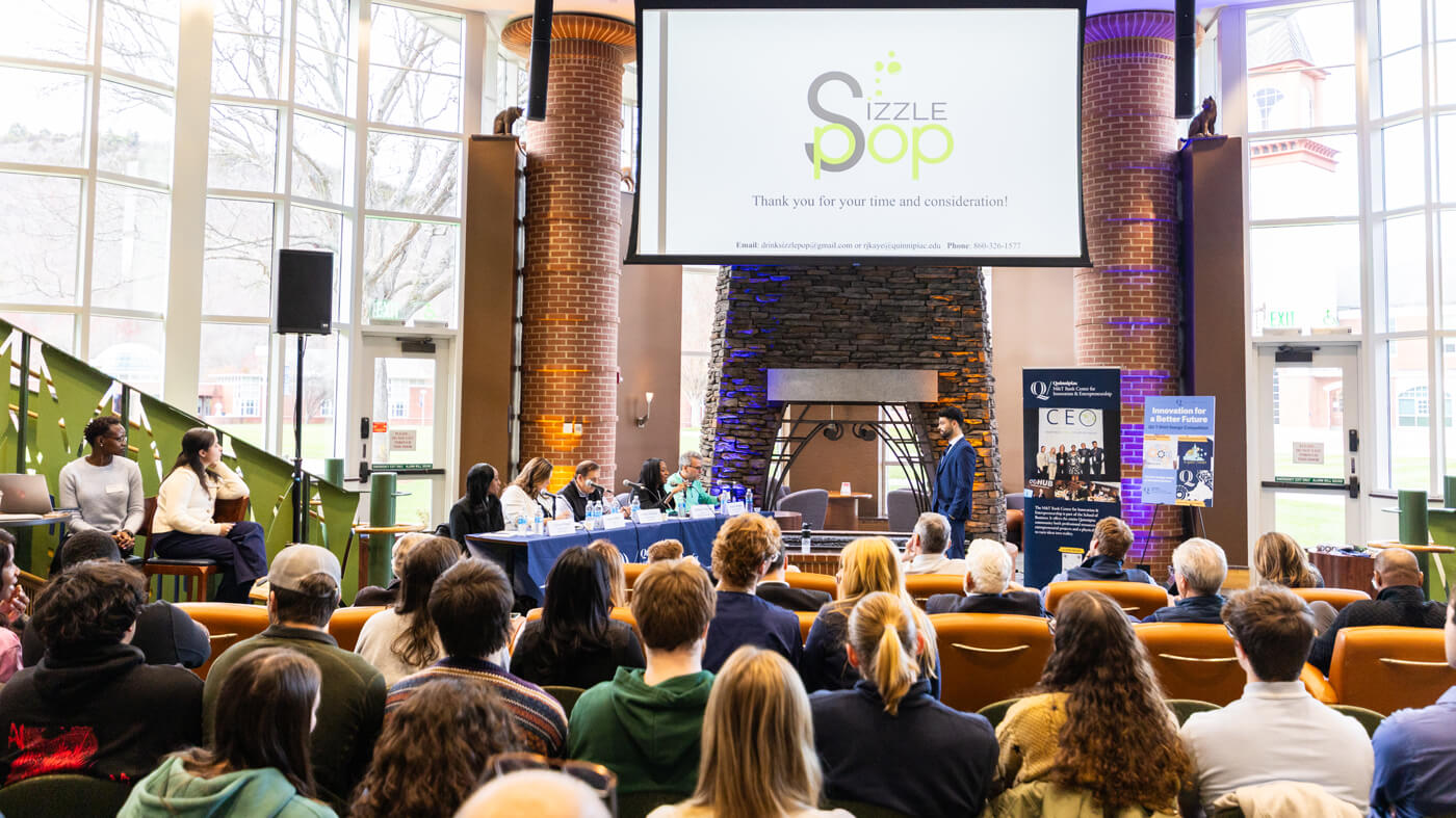 A wide shot of the pitch competition event shows a student presenting in front of a panel of judges and a full audience, with a large screen displaying the team name “Sizzle Pop.”