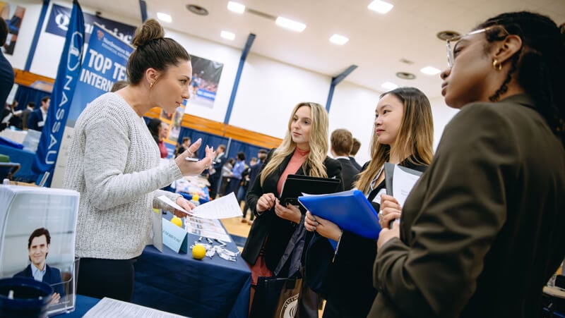 3 female students speak to a woman standing behind a career fair table