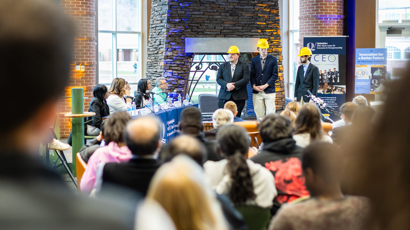 Three students wearing construction helmets deliver their pitch presentation to a packed room, standing in front of a fireplace as judges look on.