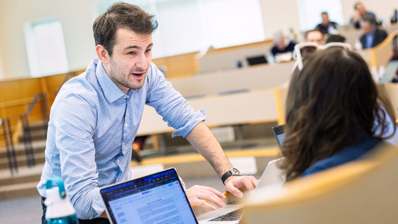 A professional gives direction to a group of students working on laptops.