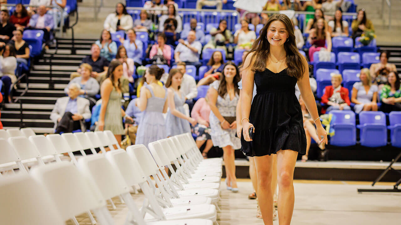 woman in a black dress leads her row into their chairs, she's smiling at the ground while she walks