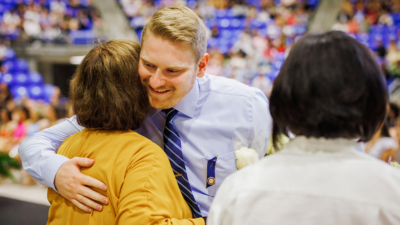 blonde male student with a beard and blue striped tie hugs the woman who presented him his pin
