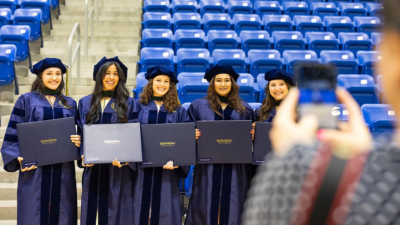 Five graduates smiling as a friend takes a group photo of them