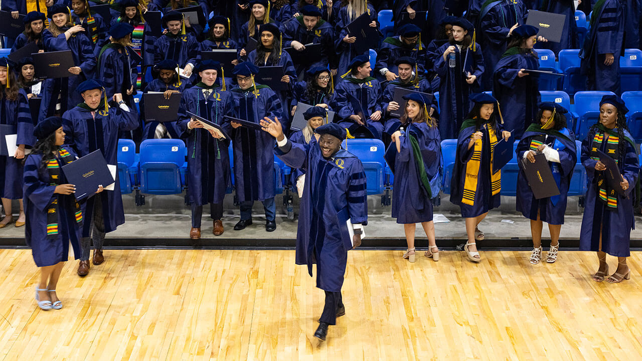 A graduate walking in front of their classmates, waving with their diploma