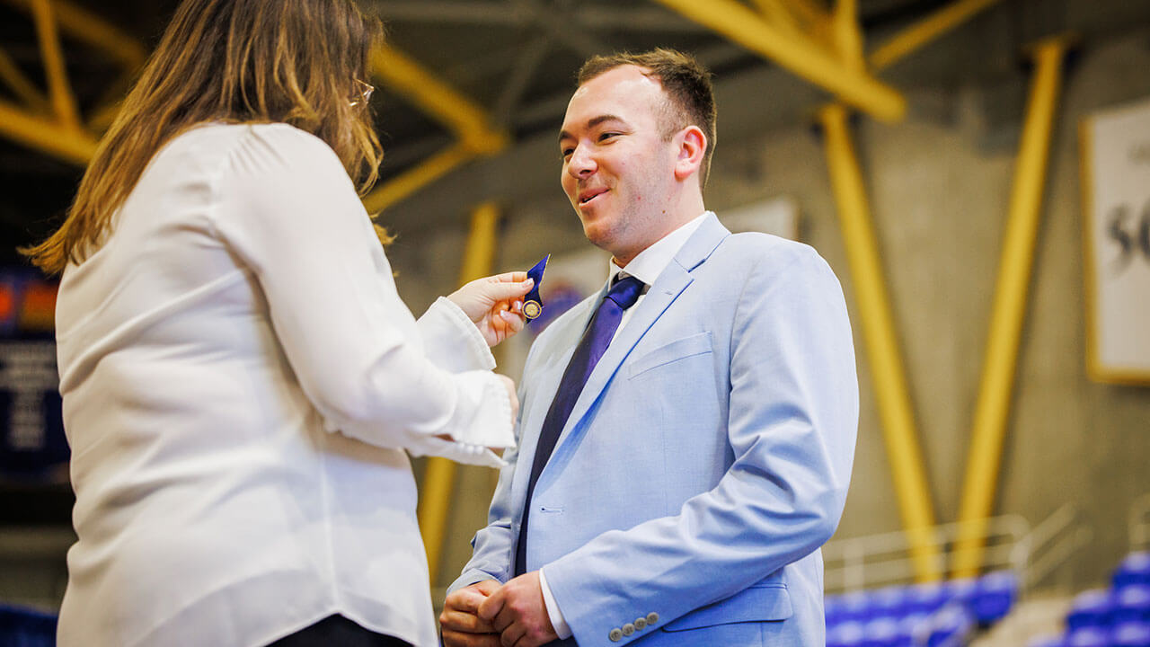 male student with brown hair in a pale blue suit and purple tie has his pin held up to his lapel before it's pinned