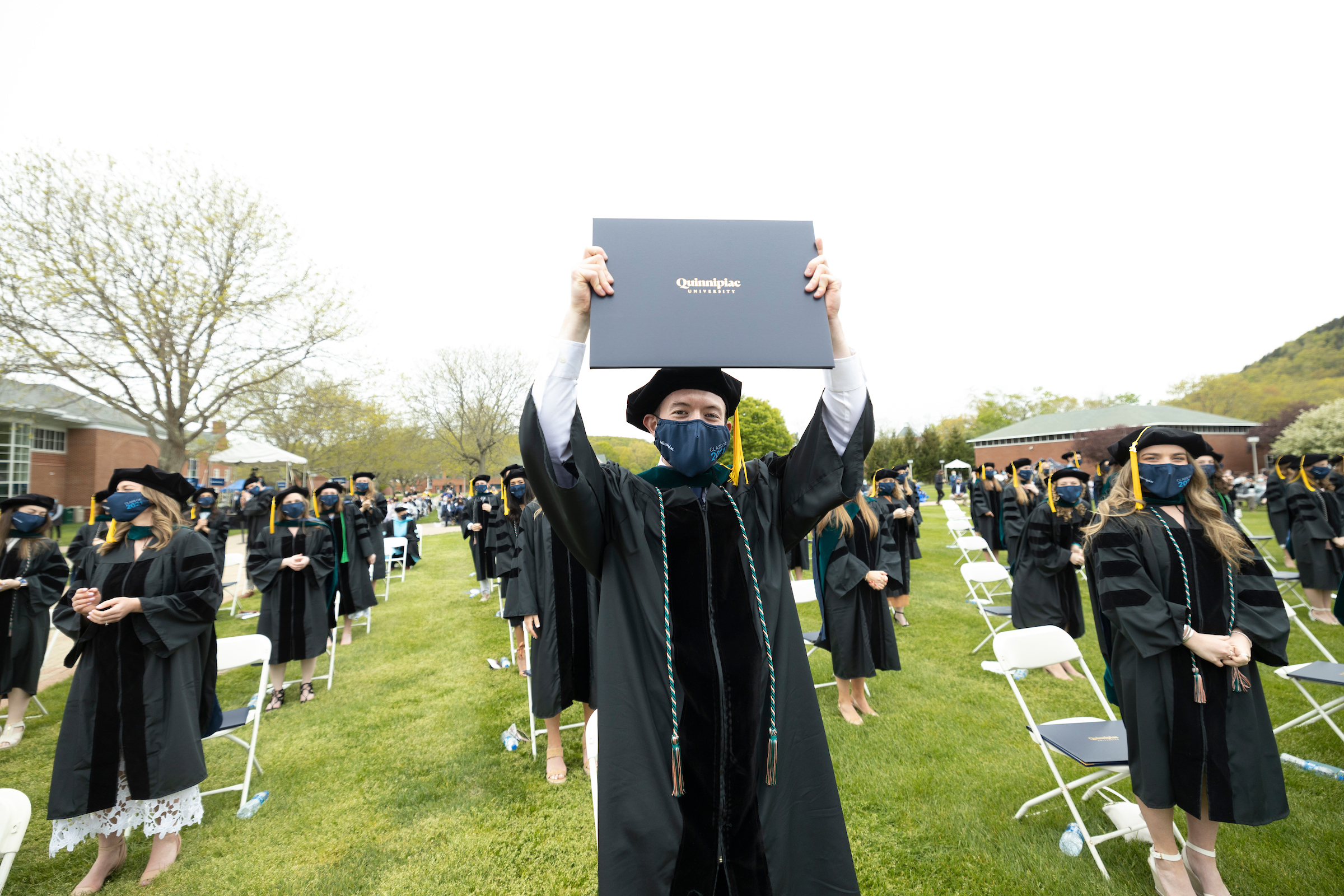 Student holding diploma in middle of quad with other students sitting for ceremony