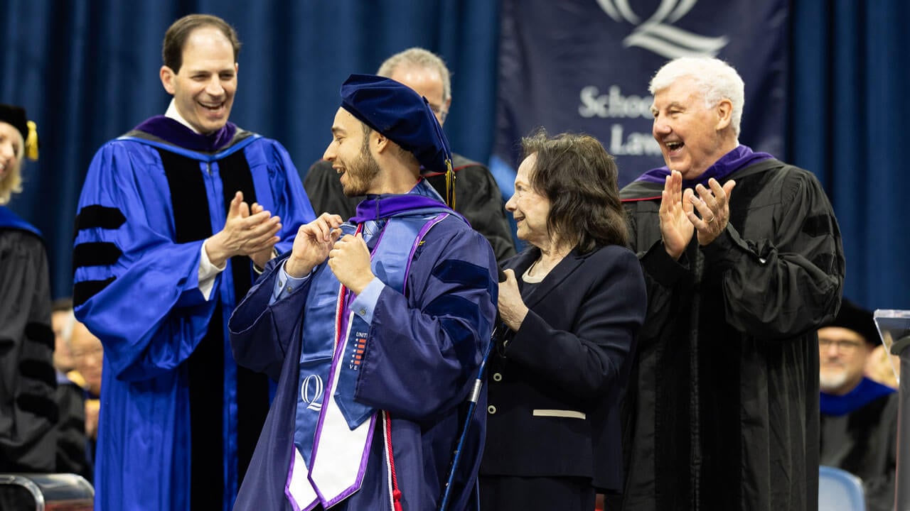 Student smiles, surrounded by professors, as they receive their hood.