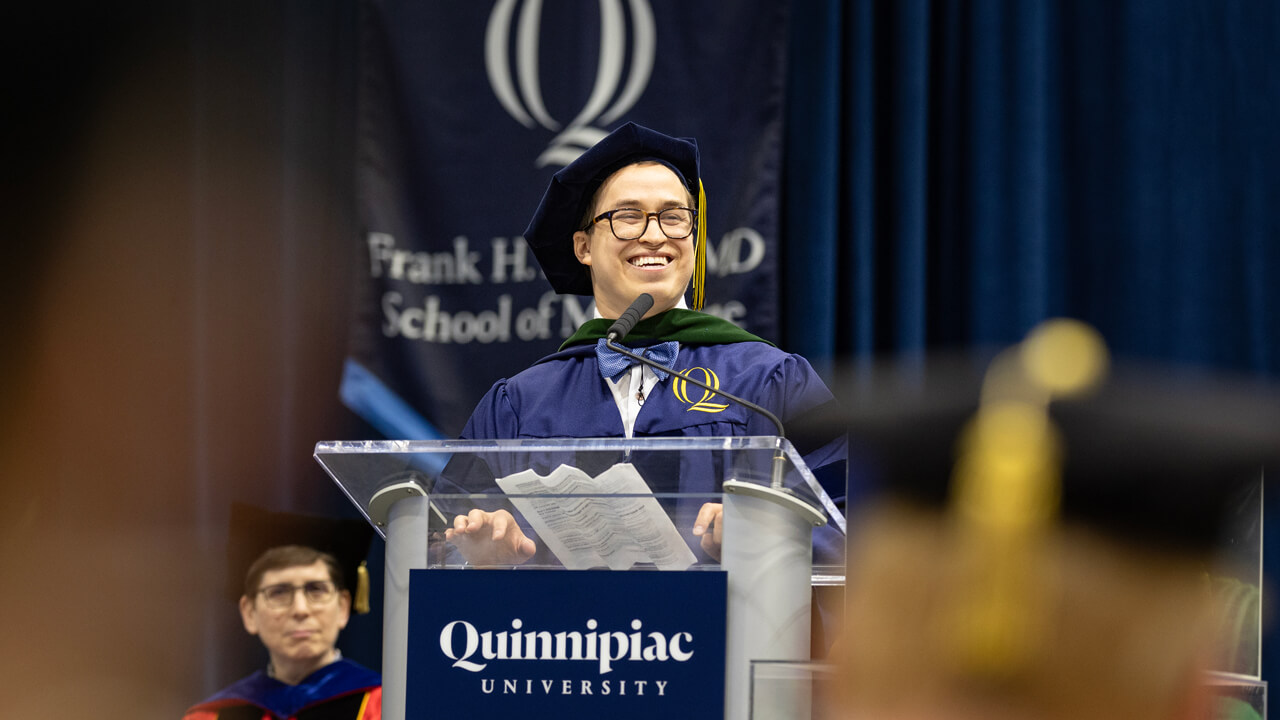 Colin Uyeki in his cap and gown smiles broadly at the Commencement podium