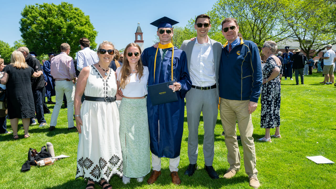 A graduate holds a diploma and smiles with 4 loved ones in front of the library clocktower
