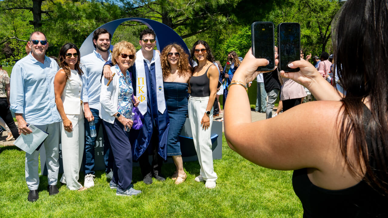 A person takes a photo of a graduate surrounded by half a dozen loved ones in front of a Quinnipiac sign