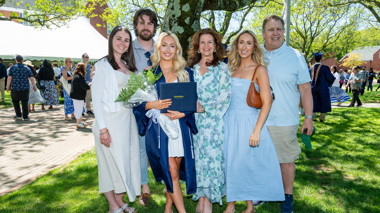 A graduate holds a bouquet and diploma and poses with loved ones under a tree for a photo