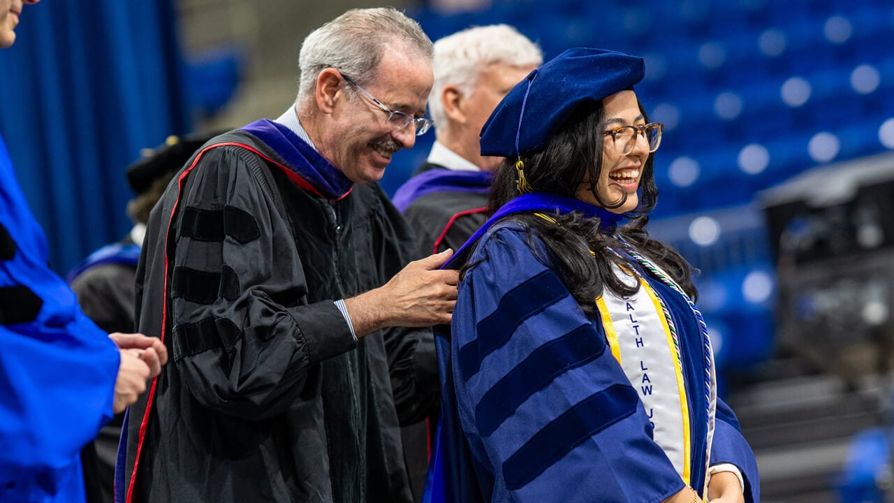 A graduate laughs as she receives a doctoral hood from a professor