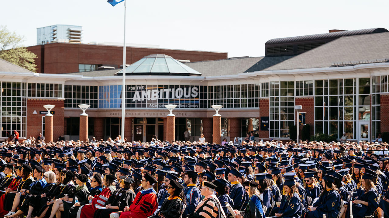A wide angle of grads sitting on the quad with the student center in the background